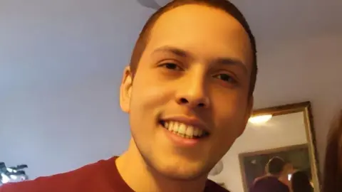Close-up photograph of Gareth looking towards the camera indoors, with a mirror visible in the background. He has cropped brown hair and is smiling.