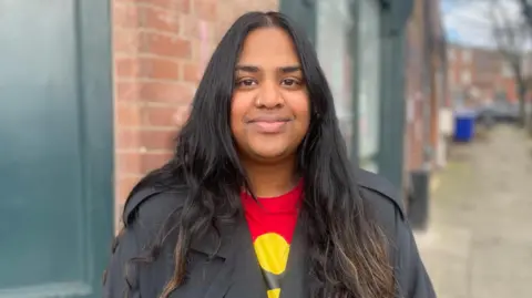 Simon Thake A woman with long dark hair and a nose piercing stands in and a grey mac over a red t-shirt