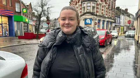 A woman stands close to the camera on a very rainy day. She has brown hair tied back in a ponytail. She is wearing a black puffer coat. The pavement is slick and reflective in the rain behind the woman, backed also by a line of traffic. Behind her is a busy street in strabane, with buildings like a subway and o2 shop in vision. 