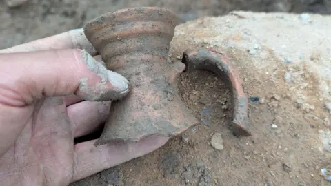 Civic A hand is holding a broken neck of a pottery flagon, with the remainder of it seen still partially buried in a trench.