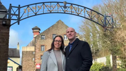 Kurt and Rebecca Canfield smiling at the camera, standing underneath the arched Jennings Brothers Castle Brewery sign. Rebecca has dark long hair and is wearing a grey coat. Kurt is bald and is wearing a black coat. Stone buildings can be seen in the background.