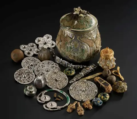 NMS A collection of metal objects, including brooches and bracelets, and a metal vessel, displayed against a black background.