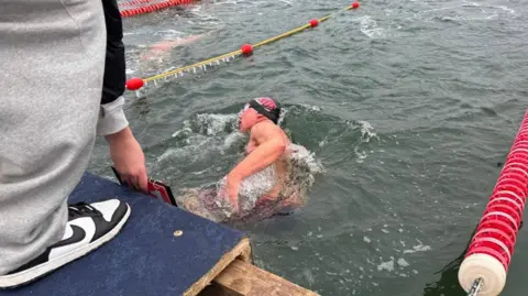 Donna Curtis Neil is swimming away from the camera wearing a dark blue swimming cap and goggles.
