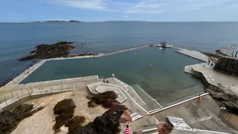 Piscinas de La Vallette en Guernsey. La piscina principal está junto al mar. Varias personas con traje de baño están en el agua o paseando por el borde.