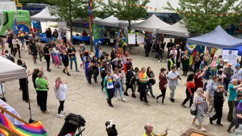 Hundreds of people walking through a pedestrianised area lined with market stalls and gazebos. Many are wearing rainbow clothes or carrying flags.