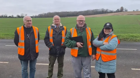 Cambridgeshire Constabulary Three men and one woman are stood at the side of a road in front of a large green field. They wear orange high visibility vests and are looking at the camera. One member is carrying a clipboard and pen.