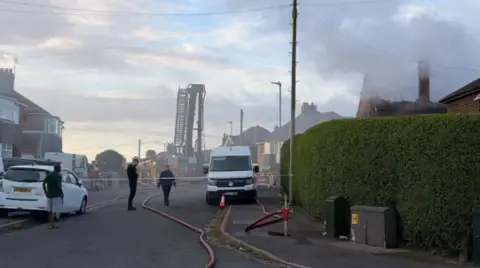 A street with vehicles parked either side and a hedgerow in the foreground. The scene is behind a police cordon, with a fire engine and ladder visible through smoke. A house on the right hand side of the image is smouldering.