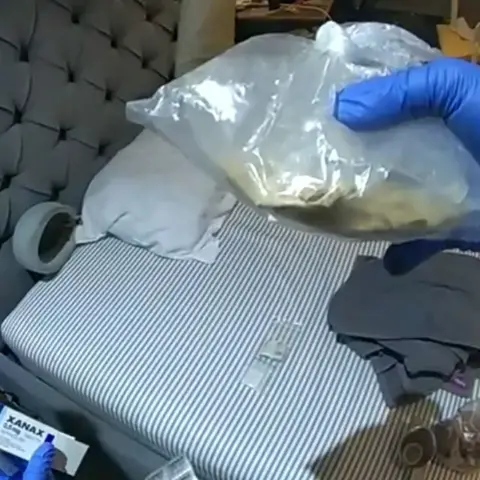 A police picture inside the flat of Metropolitan Police Zo. A bed can be seen and an officer's hand is holding a plastic bag with a substance.