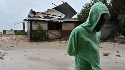 EPA/Shutterstock A man walks by a house damaged by the preliminary winds of Hurricane Melissa