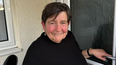 Ben Schofield/BBC Anita Wright at the front door of her home. She is smiling and looking directly down the camera. She has blue eyes, short dark brown hair and is wearing a black top. One of her hands is resting on the handle of her front door, which is in the right hand bottom corner of the picture.