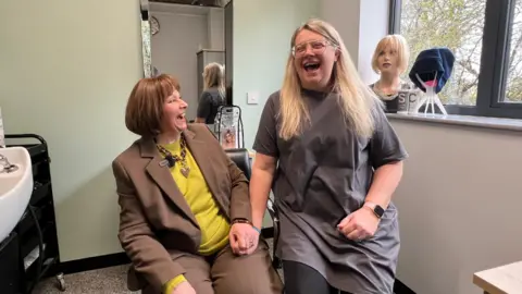 Two women sat next to each other in a consultation room laughing and holding hands