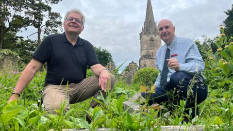 Paul Frowde and Julian Carosi crouched in the graveyard at St Bartholomew's Church with the church steeple behind them
