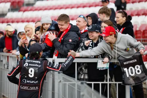 TŻ Ostrovia/Wojciech Tarchalski A speedway rider wearing a black racing suit with the number 9 on the back reaches over a barrier to greet spectators in the stands. The fans are leaning forward with scarves, phones and things to be autographed.