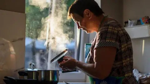 A woman cooks at a hob. She has on a checked, half-sleeve top and holds a saucepan lid in her hands. On the hob are two pots, one covered, one without a cover and behind her is a window showing trees outside