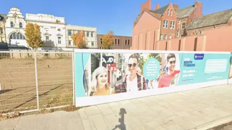 Google An area of cleared ground behind temporary fencing in South Shields town centre. It is the site of the planned new Tyne Coast College campus. Promotional words for the scheme sit alongside an image of three smiling, generic promo style, young people. This is plastered on the fencing like a billboard advert. There are a few trees at the far side of the cleared ground in front of buildings along King Street. It is very sunny with no clouds in the sky.
