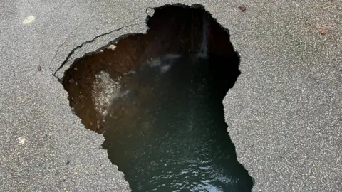 Nicola Haseler/BBC A large hole in the middle of an asphalt road, exposing a deep cavity filled with water .