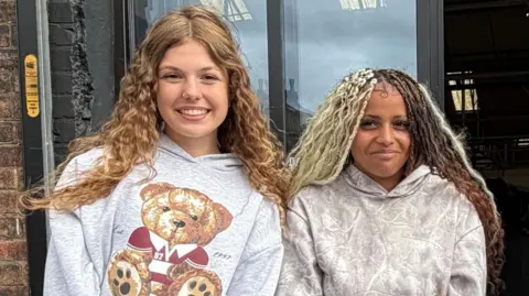 A girl with long curly blonde hair, wearing a grey hooded top with a picture of a teddy bear, stands next to a girl with blonde and brown curly hair, who is also wearing a grey hooded top. Both are smiling as they stand outside the front door of a hall.