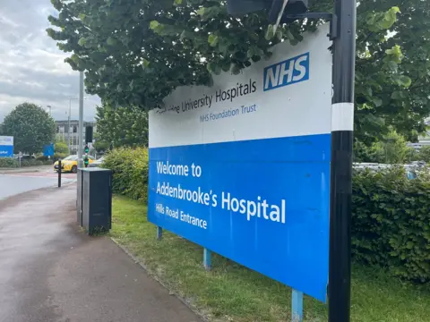 Jozef Hall/BBC The blue and white Welcome sign at Addenbrooke's Hospital, next to a hedge and under a tree. In the distance is a car park and the hospital building.
