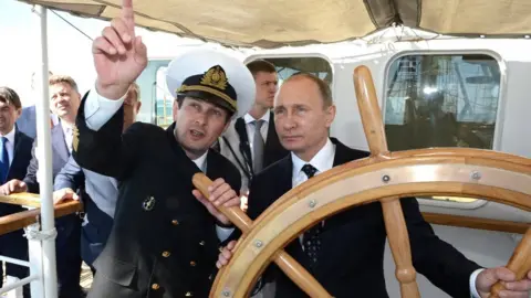 Russian government Vladimir Putin is seen at the helm of a boat as an officer points out something to him (September 2016)