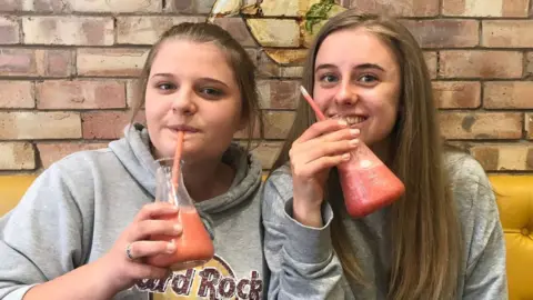 Family photo Gracie and Laura Nuttall sitting together, both drinking from a glass with a red-orange liquid in it that looks like a scientist's conical flash. Gracie is on the left and is wearing a grey Hard Rock Cafe hooded top while Laura is on the right and is wearing a long-sleeve grey jumper