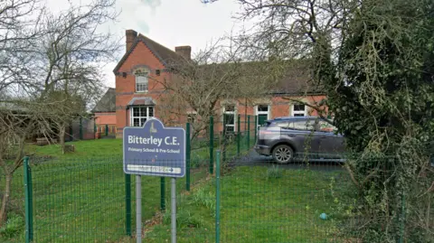 A sign which reads "Bitterley C.E. Primary School and Pre-School" outside a school building. With a car parked on the right hand side and grass surrounding the building.