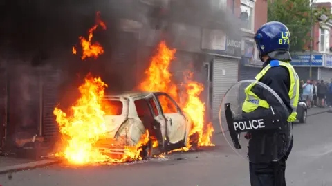 A police officer holding a large round plastic riot shield and wearing a helmet looks on as a car burns.