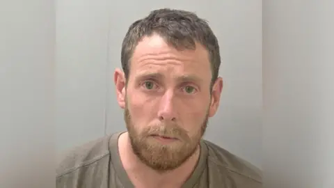 A police mugshot of a man with short brown hair, moustache and beard. He is wearing a brown T-shirt and looking at the camera.