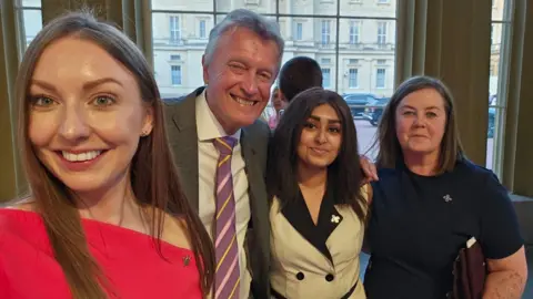 Four people stand in a line smiling at the camera, one of whom, a woman on the left, who is taking a selfie. She has long brown hair and is wearing a red dress. A man with grey hair, wearing a white shirt and purple- and yellow-striped tie under a grey suit jacket, is standing slightly behind her. He has his arm around a woman with long dark hair, wearing a cream and black dress. On the end is a woman with shoulder-length dark hair who is wearing a navy blue t-shirt.