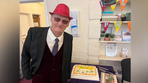 Lily-May Symonds/BBC Pat poses for the camera next to his cake, which is a large white rectangular cake with orange and yellow decorations on and white icing. Pat wears a red sparkly hat, sunglasses, and a three-piece suit with blue tie and white shirt.