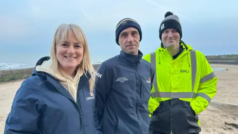 Sarah, who has long blonde hair, Carl, and Neil, who is taller and clean shaven, stand by side. All are are wearing RNLI coats.