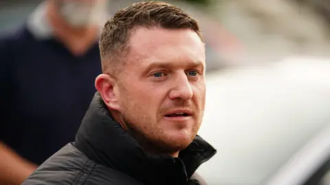 PA Media Tommy Robinson looking to the right front of the photo. He is wearing a black jacket with the collar up. He has light stubble and short cropped auburn hair.