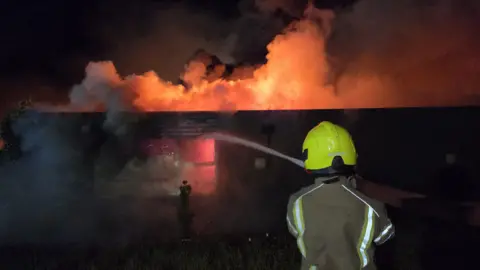 Suffolk Fire and Rescue Service The back of a firefighter in the foreground holding a hose with a jet of water aimed at a smoking building during the night. The building is giving off an orange glow