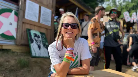 Lucinda Tait sat at a table in Strummerville, wearing sunglasses, a white T-shirt with green stripes and colourful festival wristbands.