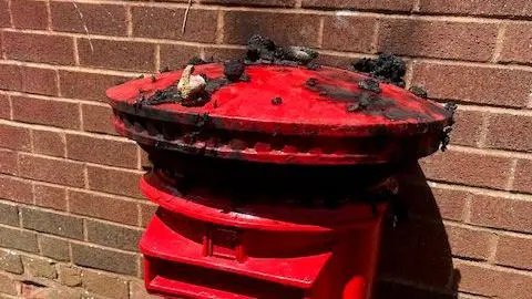 A red post box with a brown brick wall behind it. The tope of the postbox looks burned and there are bits of burned fabric on the top. 