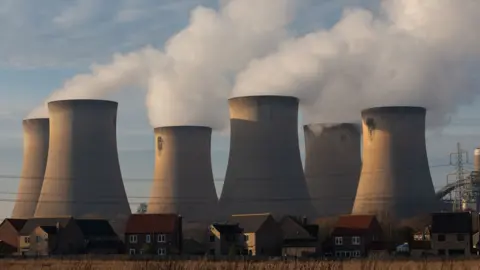 Draw getty image power station. The photo features smoke billing from six stout chimney. There are a row of houses and several electric cables in the foreground.