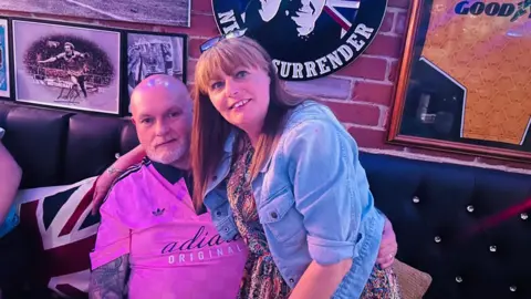 A photo of a man and a woman smiling together. The man is bald and is wearing a pink top. The woman has ginger hair with a fringe and is wearing a blue denim jacket. 