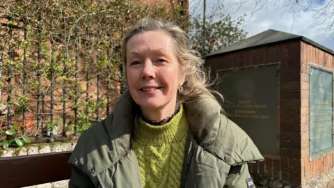 A blonde-haired woman wearing a green jumper and green coat, standing in front of a brick pillar and raised railings covered in vines.