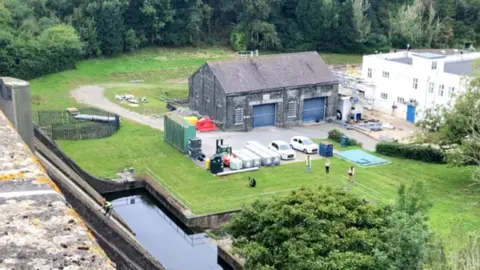 A stone building with two large blue garage-like doors. The building is next to the edge of a reservoir and is surrounded by grass.