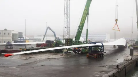 A large single wind turbine blade is lifted by crane onto specially designed trucks on a foggy dockside