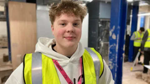 A teenage boy with light brown curly hair is standing in a construction environment. He is wearing a high-vis jacket over a white hooded top and in the background are other students and tools.