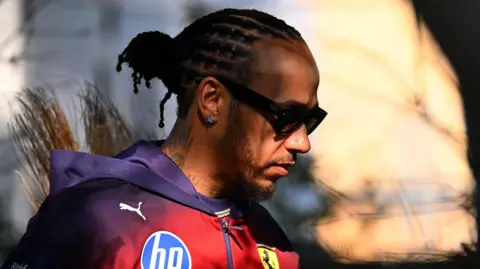 Getty Images Sir Lewis Hamilton wears a Ferrari jacket as he steps into a dappled sunlight. He is wearing sunglasses.