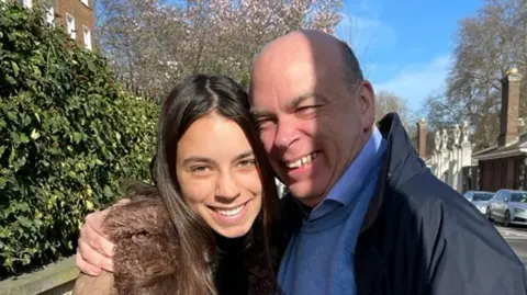 PA Media Hannah and Mike Lynch hugging and smiling into the camera. Miss Lynch has long brown hair and a brown coat. Mr Lynch has a blue jumper and shirt collar, and a black coat.