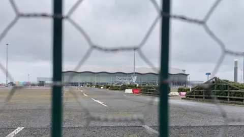 BBC/Victoria Scheer Doncaster Sheffield Airport terminal building surrounded by empty carparks. The picture is taken through a wire fence. 