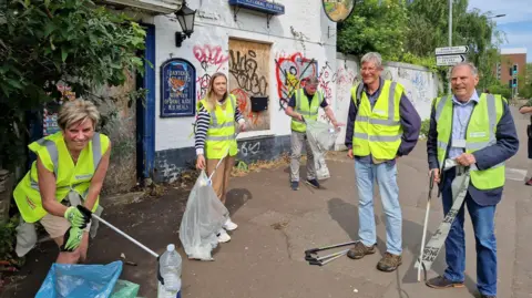 Five people holding litter pickers and bags standing in front of a pub. They are wearing hi-vis jackets and smiling