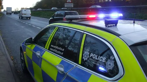 Suffolk Police Police car with flashing lights parked on a dual carriageway with cars driving past on either side.