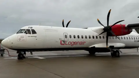 A white passenger aircraft is parked on the runway at Jersey Airport. It has the logo Loganair painted along its side in red.
