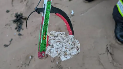 A man using a litter picker holds a white, waxy lump while standing on a sandy beach