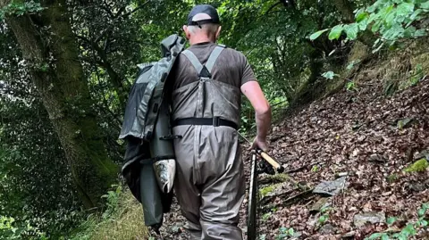 Natural Resources Wales Stephen Samuel, wearing dungarees, is seen from behind with a whole salmon protruding from the sleeve of a coat slung over his shoulder