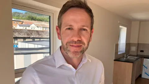 A man with short brown hair and stubble on his face wearing a white shirt smiles while standing in a kitchen.