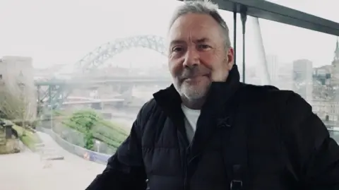 BBC Andrew Parker is standing in front of a large window overlooking the Tyne Bridge. He is wearing a white t-shirt and a black puffer jacket. He has grey hair and beard and is carrying a backpack. 
